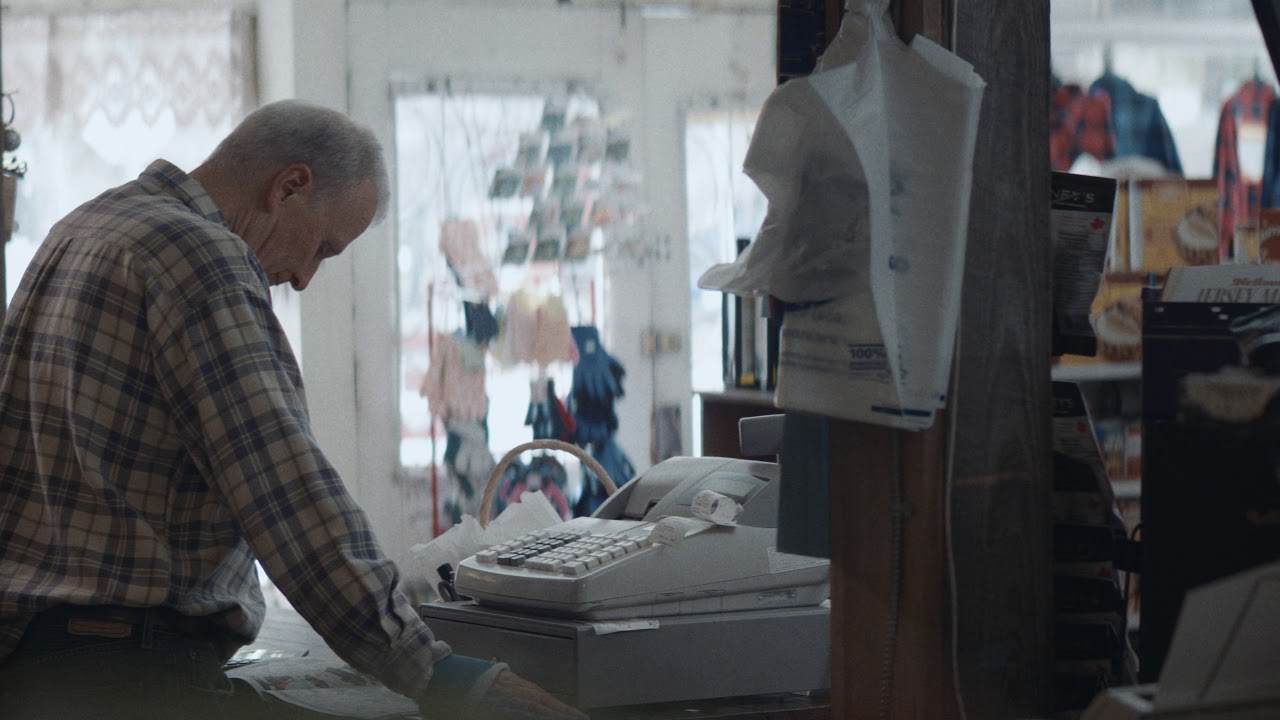 An older shopkeeper stands behind the counter of a small general store.